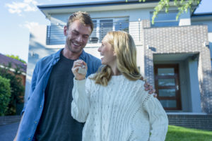 Young couple holding their new house key. They are standing in front of the home in casual clothes. Both are happy, relaxed and smiling. He has a beard and she is blonde. They could be buying or selling real estate. Copy space